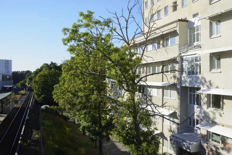 Boendets fasad sedd från ett fönster på tredje våningen. Framför boendet står rett träd och till vänster syns den närliggande tunnelbanestationen.