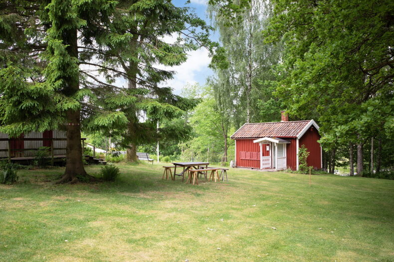 En närliggande gräsyta med stora träd, träbänkar och ett bord att sitta vid, samt en stuga lite längre bort.