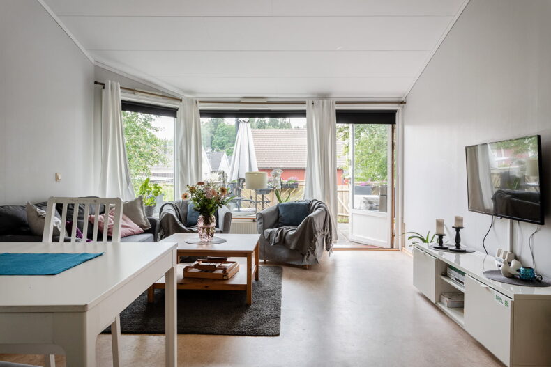 Ett rum med grå soffgrupp, soffbord och TV i förgrunden samt stora fönster och en altandörr i bakgrunden.