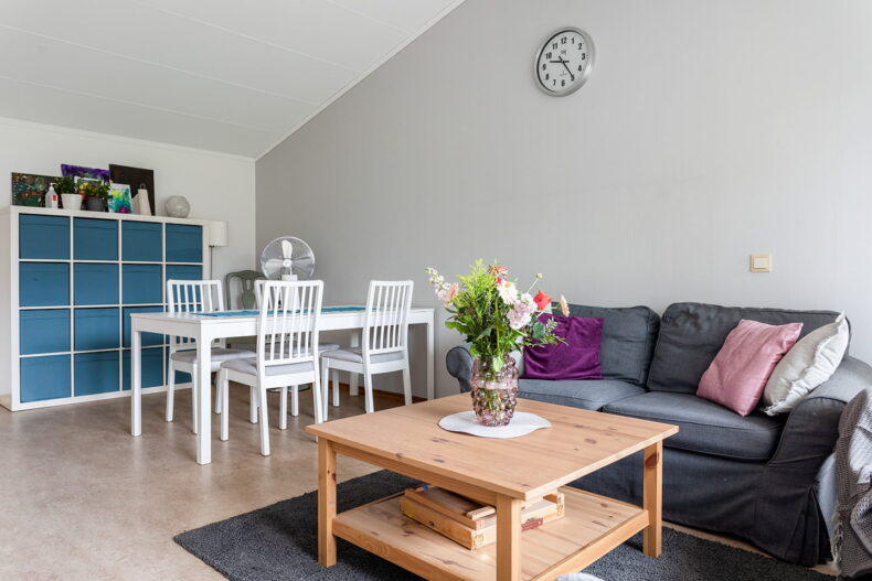 Ett rum med snedtak. Till höger står en soffa och ett soffbord och till vänster ett matplats och en hylla med blå lådor i och konst ovanpå.