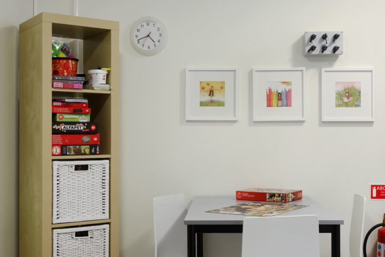 En brädspelshörna med flera spel i en hylla bredvid ett bord som det ligger ett pussel på. På väggen hänger små tavlor och en klocka.