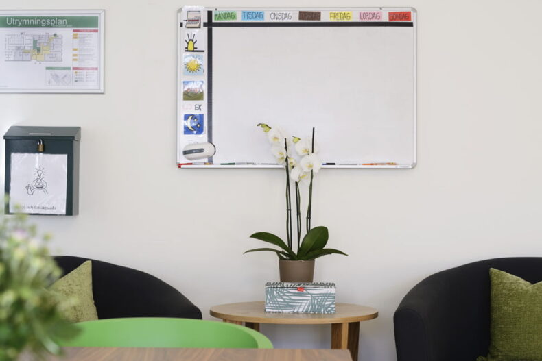 På en vägg hänger en whiteboard med veckoplanering och bildstöd. Nedanför står ett litet bord med en orkidé.