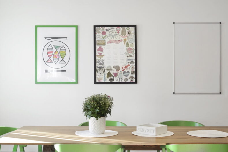 Ett långbord med gröna stolar och en krukväxt. På väggen hänger tavlor med mat-tema.