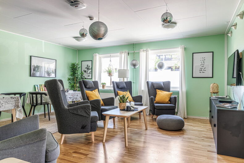 Gemensamt utrymme med gråa fåtöljer, gula kuddar och TV.