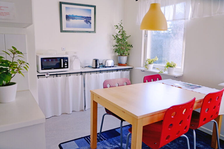 köksbord, röda stolar, mikro, tekokare på hylla mot vägg, gul lampa fönster