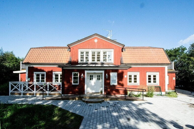 Boendet sett framifrån, stor röd villa med vita knutar, ramp till entrén samt stenläggning runt omkring.