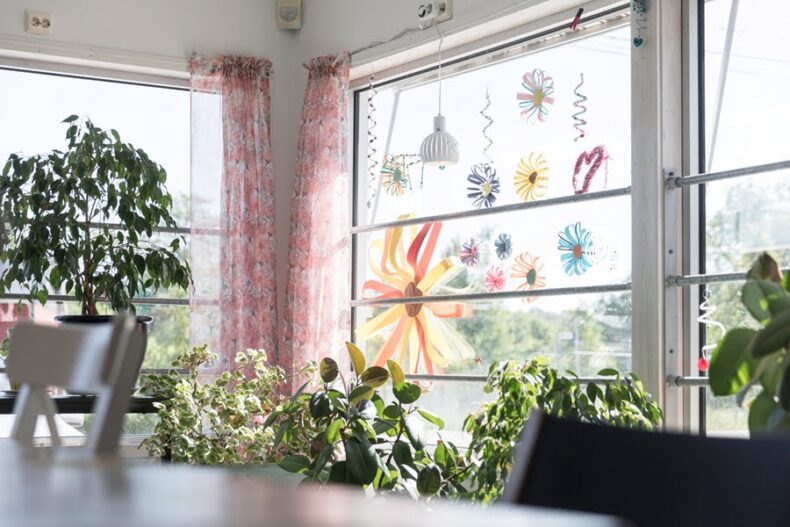Många krukväxter framför ett fönster som det hänger flera pappersblommor i.