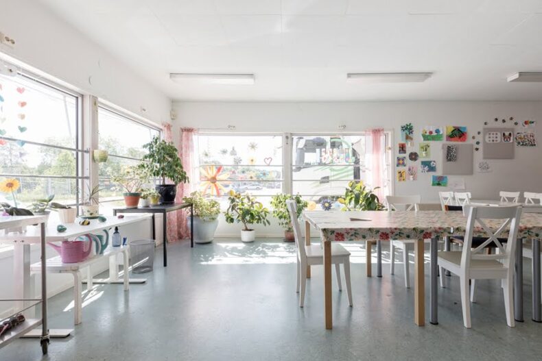 Ett ljust rum med ett långbord i mitten. Väggarna har stora fönster och det står många krukväxter på bord och golv framför dem