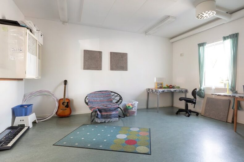 Ett rum med en låg korgstol, ett litet skrivbord, och ljust fönster. På golvet står en gitarr och ett elpiano.