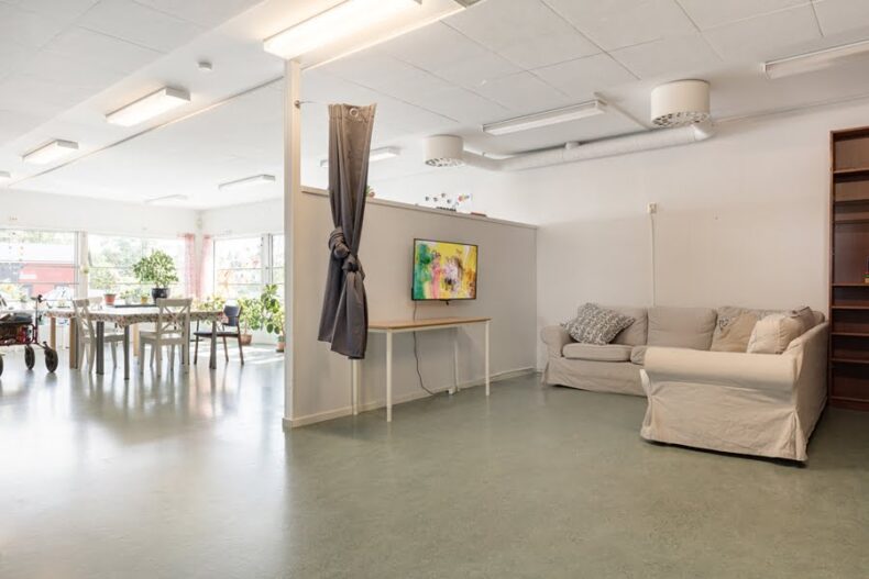 En TV-hörna med soffor i förgrunden med en skiljevägg mot ett ljust rum med långbord och många krukväxter i bakgrunden