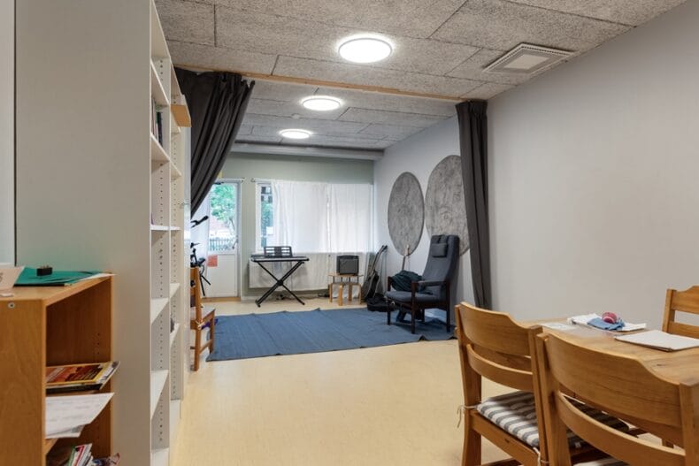 Ett rum som går att dela av med en gardin i mitten. I den närmare delen finns ett bord och en förvaringshylla, och i den bortre delen finns en fåtölj och ett elpiano