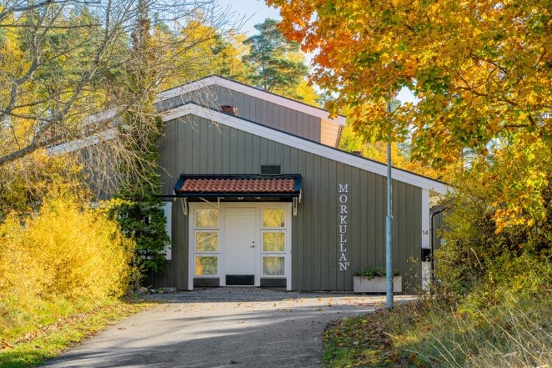 Boendets entré och grå fasad med träd och buskar runtom