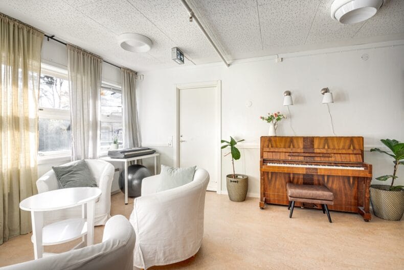 Ett ljust rum med fåtöljer runt ett litet bord vid fönstren till vänster samt ett piano till höger