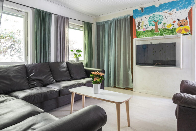 Tv-rum med svarta soffor, stora fönster med gröna gardiner. Stor tavla på väggen målad av barn