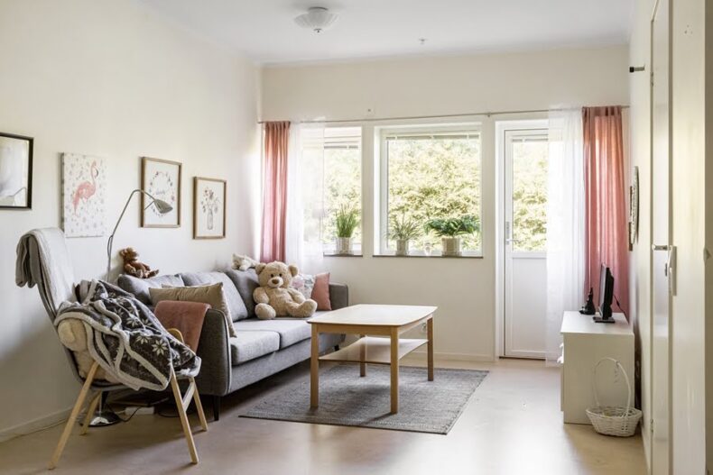 Vardagsrummet i en av boendets lägenheter, med soffa, soffbord, TV och dörr till uteplats. I soffan finns en stor nalle och på väggen hänger flera tavlor.