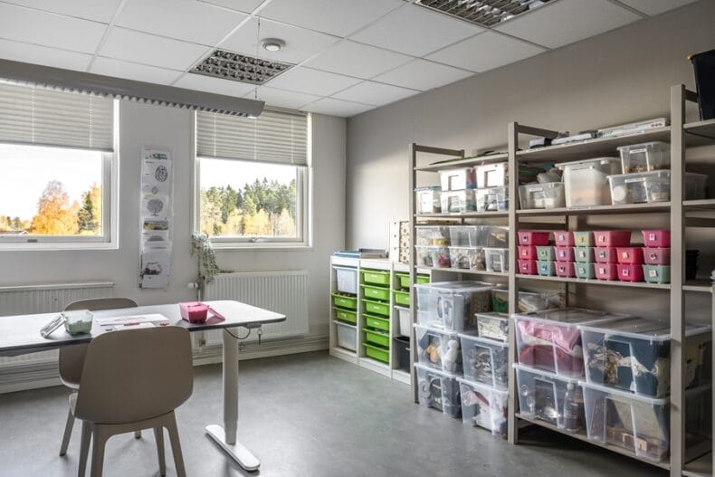 Ett rum med ett bord i mitten och flera hyllor vid väggen med material för skapande