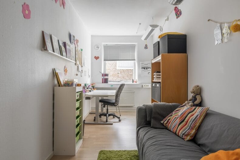 Ett avlångt rum med soffa och en liten hylla i förgrunden samt ett skrivbord och en större hylla längre bort.