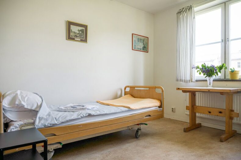 Ett av boendets rum med höj- och sänkbar säng, ett litet bord med en blomvas på, tavlor på väggen och ett ljust fönster