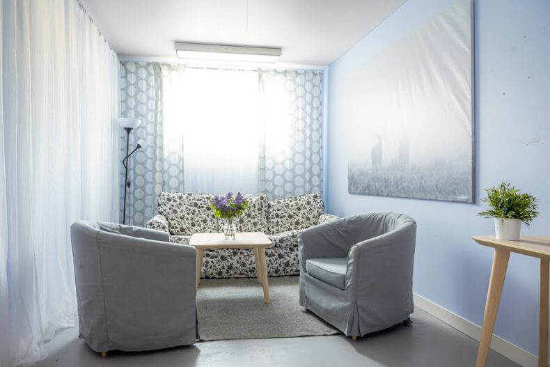 Ett litet rum med två fåtöljer, en soffa och ett soffbord med en blomvas. Framför ena väggen samt fönstret hänger skira gardiner som sprider ljuset i rummet.