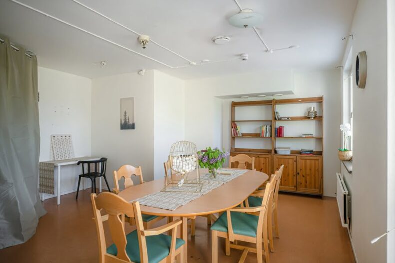 Ett gemensamt rum med ett långt bord och stolar i mitten. I bakgrunden står en bokhylla och ett litet skrivbord.