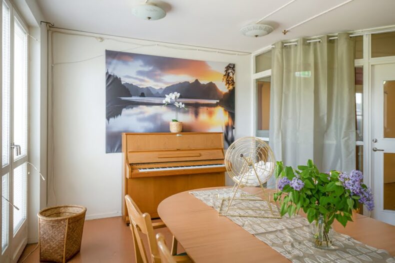 Ett gemensamt rum med ett bord och stolar i förgrunden samt ett piano i bakgrunden. På väggen hänger en stor landskapsbild och det hänger draperier mot fönstren till den intilliggande korridoren.