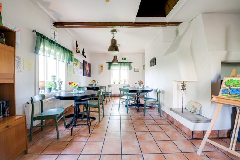 En fikalokal i en äldre byggnad med en gammal öppen spis, ett staffli i förgrunden och mycket konst på ena väggen.