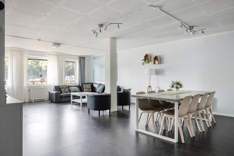 Gemensamt luftigt rum med matbord i förgrunden, soffa, fåtöljer och soffbord vid fönstren i bakgrunden