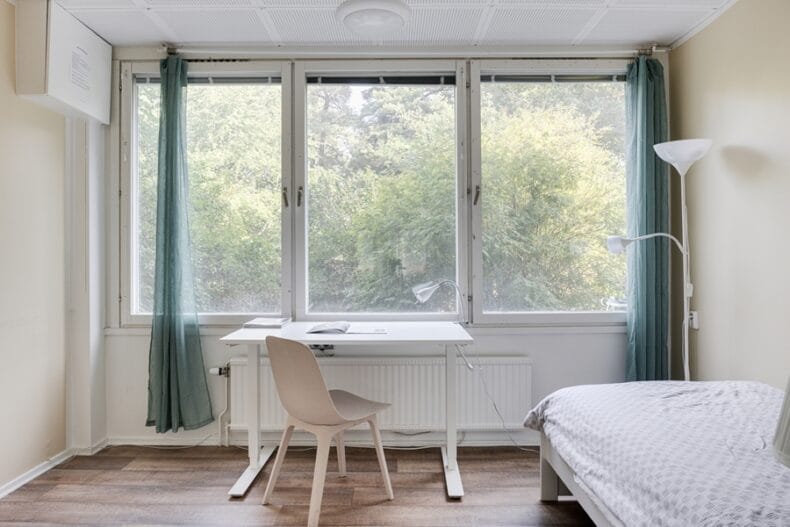 Sovrum med säng och skrivbord. Utanför de stora fönstren växer ett grönt buskage