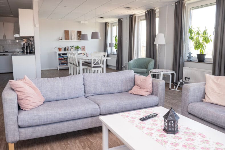 Soffor i förgrunden och kök med matplats i bakgrunden. Stora ljusa fönster längst väggen till höger.