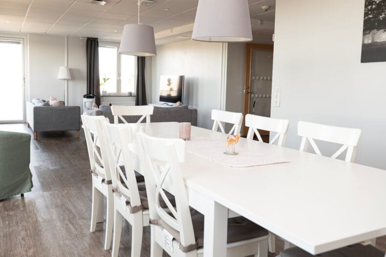 Vitt stort matbord med soffgrupp i bakgrunden och stora fönster till vänster.