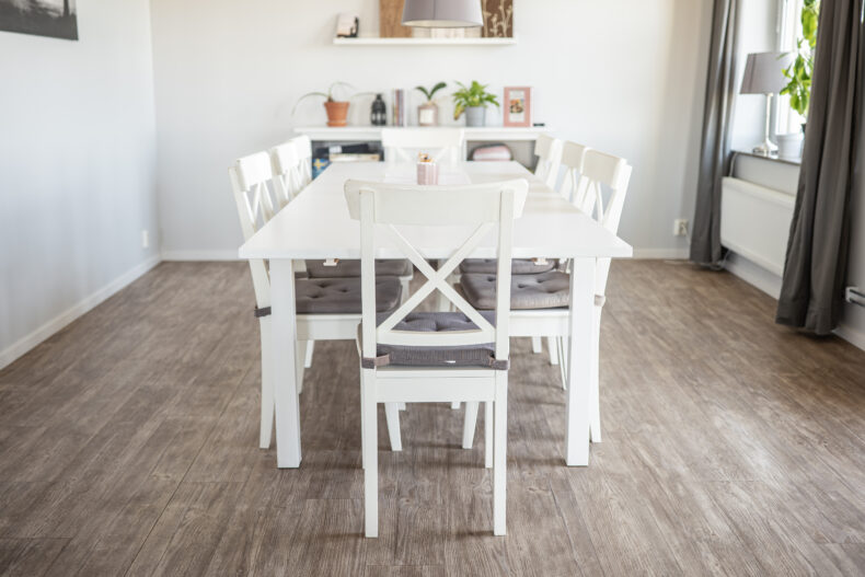 Stort vitt matbord framför hylla med sällskapsspel. Till höger stora fönster med grå gardiner.