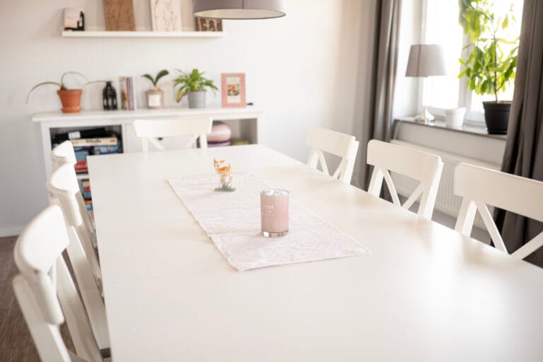 Stort vitStort vitt matbord framför hylla med sällskapsspel. Till höger stora fönster med grå gardiner.