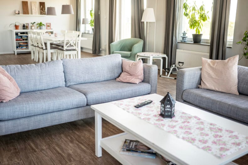 Soffor i förgrunden och kök med matplats i bakgrunden. Stora ljusa fönster längst väggen till höger.