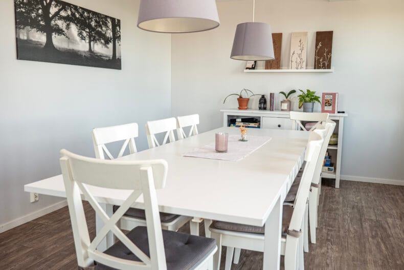Stort vitt matbord framför hylla med sällskapsspel. Till höger stora fönster med grå gardiner.