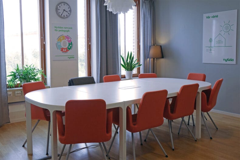 Konferensrum med stort vitt ovalt bord och sju orange stolar. Grå väggar och stora fönster med grå gardiner