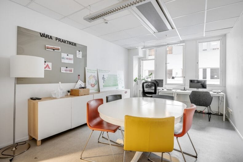 Rum med stort runt bord och fem stolar i mitten, två skrivbord med datorer vid fönstren längre bort, lång byrå till vänster.