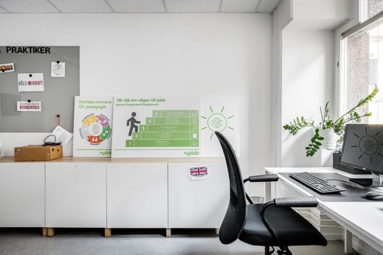 Skrivbord med dator vid ett fönster till höger, skåp och bilder på Nytidas jobbtrappa och ramverk för pedagogik i mitten och till vänster