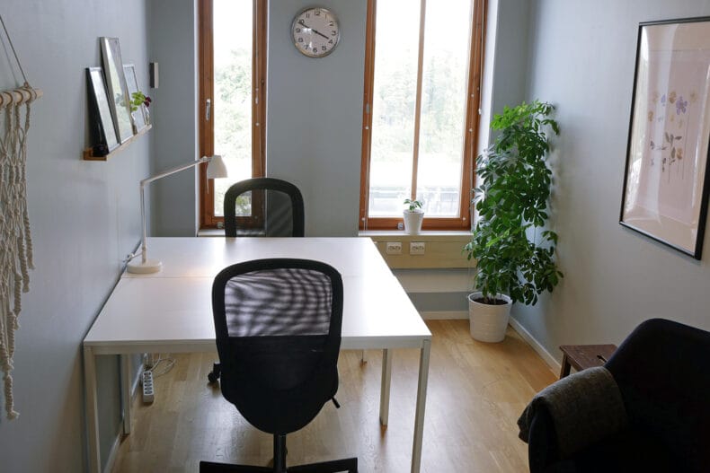 Litet rum med ett vitt skrivbord och två svarta stolar. Växt i hörnet och svart fåtölj i förgrunden