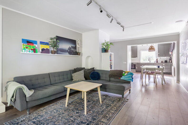 Tydlig bild på soffan, tavlor som står ovanför. Köket syns i bakgrunden