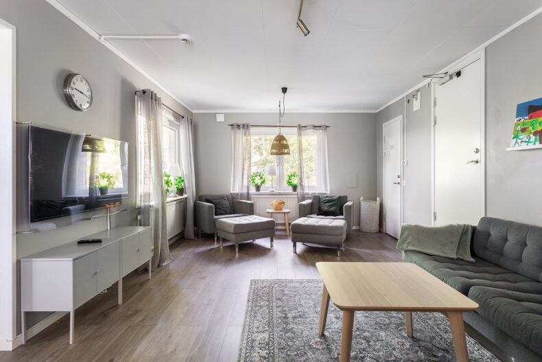 Gemensamt utrymme med soffa, mönstrad matta och tv mittemot. Fåtöljer i bakgrunden intill fönstren