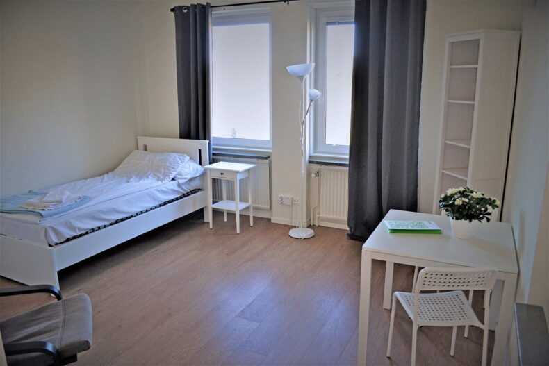 Ett av sovrummen på boendet, med säng, skrivbord, bokhylla, fåtölj och två fönster.