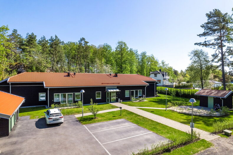 Boendet sett från en närliggande höjd, med en stenlagd gång framför entrén och skog i bakgrunden