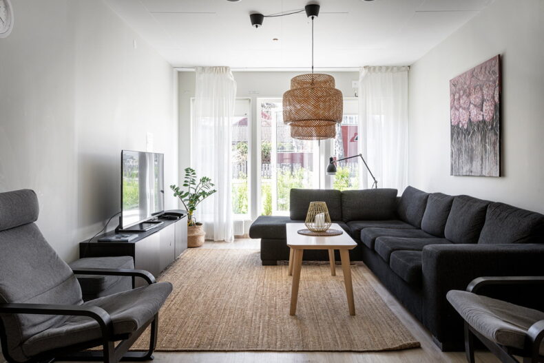 Boendets gemensamma vardagsrum med stor soffa, fåtöljer, soffbord, TV och stora ljusa fönster med ljusa gardiner framför