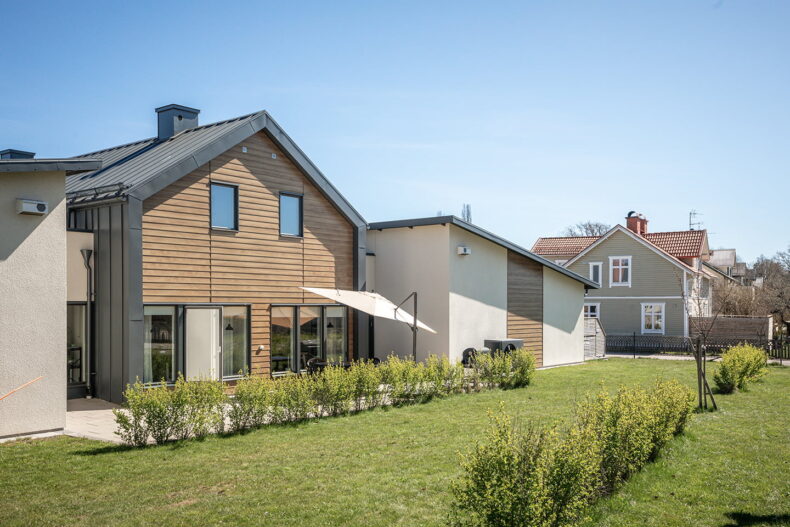 Boendets brunsvarta fasad i två våningar, med gräsmatta och nyplanterad häck framför.