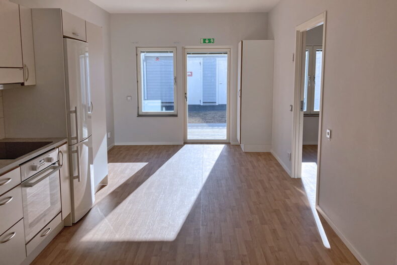 En oinredd lägenhet på boendet, med kök och vardagsrum i öppen planlösing och öppen dörr till uteplatsen