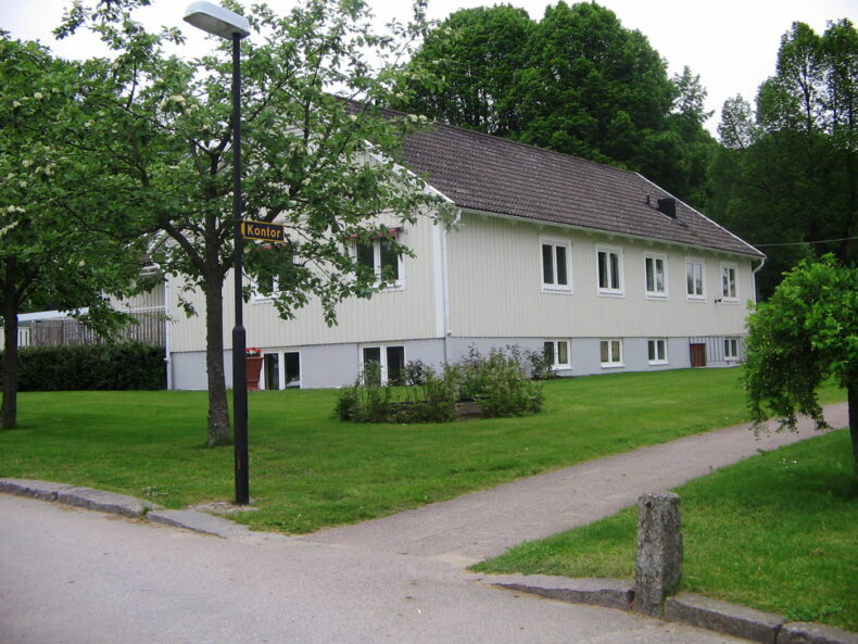 Boendets fasad, vit byggnad i två plan med källare. Gräsmatta och träd framför.