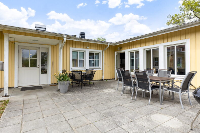 Boendets stenlagda uteplats med två grupper utemöbler samt blommor i en stor kruka vid ena dörren