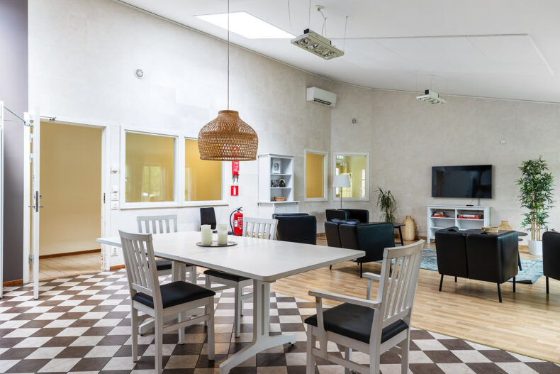 Ett gemensamt rum med matplats, fåtöljer samt TV. Det finns fönster till korridoren på andra sidan ena väggen.