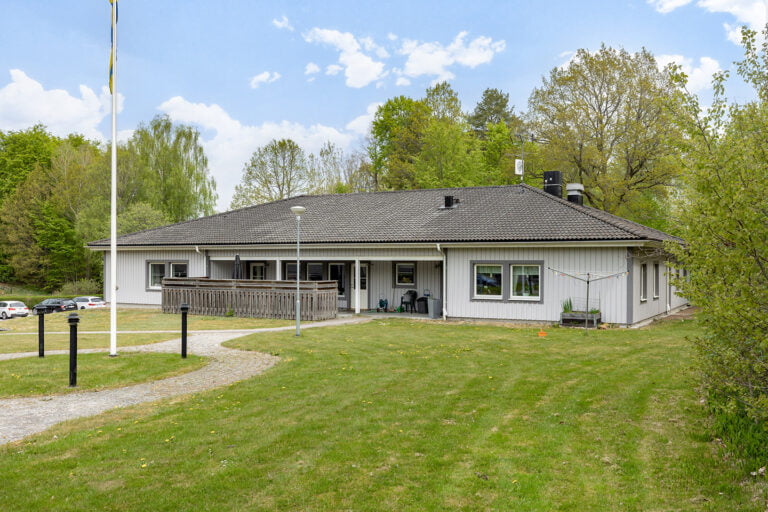 Boendets fasad, en ljusgrå enplansvilla med flaggstång, grusväg och gräsmatta framför.Ljus fasad Skogsgatan 7