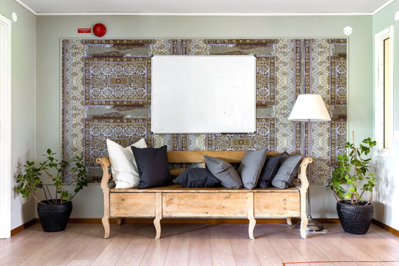 Närbild på gammal kökssoffa med många kuddar. Krukväxter bredvid och whiteboard på den mönstrade väggen bakom.